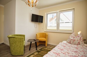 A bedroom with a bed, a green armchair, a yellow armchair, a small table, a TV mounted on the wall, and a window with a view of buildings. - Ferienhaus Bauernrose - Die Ausstattung vom Ferienhaus Bauernrose (Trent)
