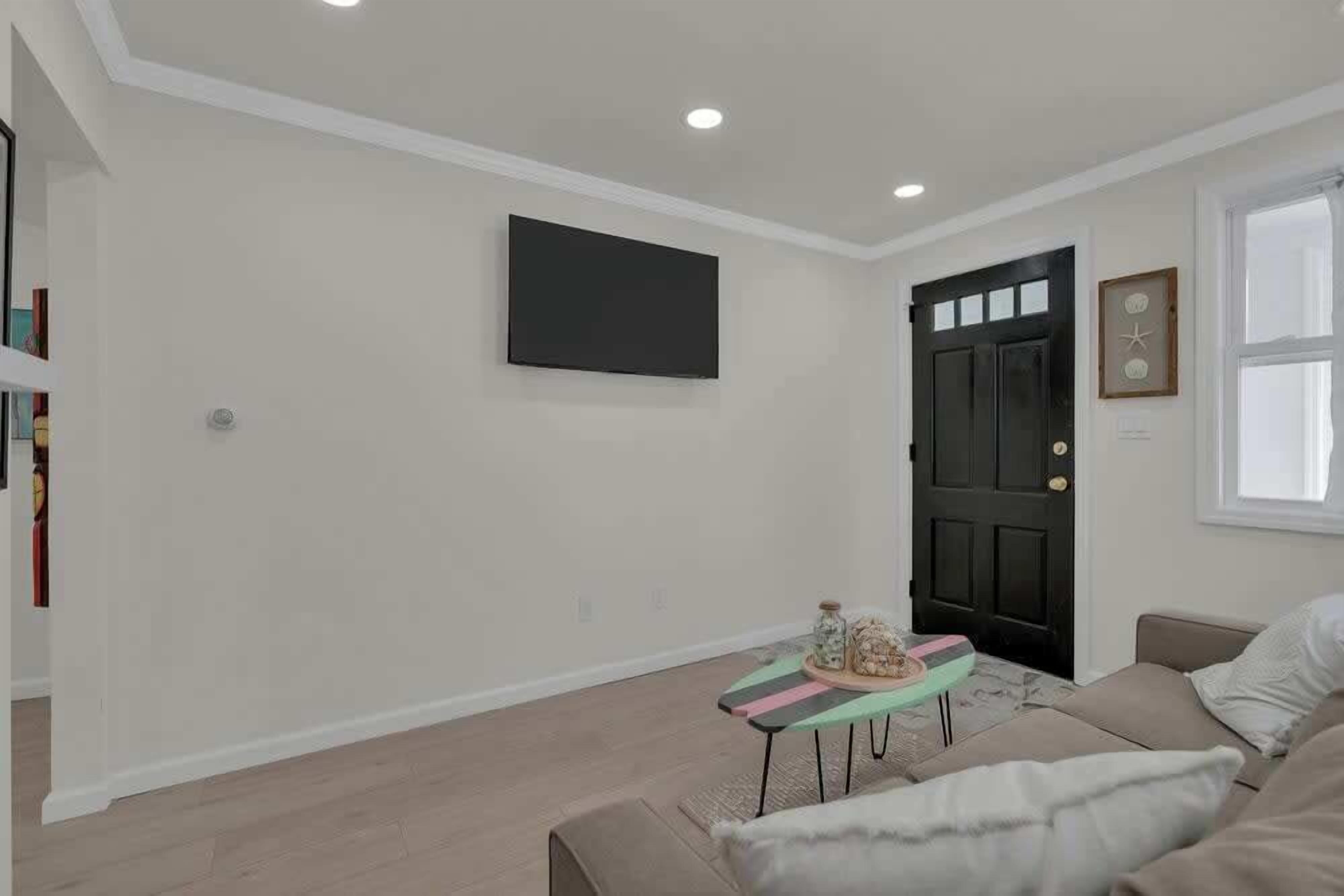 Un salon avec un canapé beige, une petite table ornée de quelques objets décoratifs, une porte noire et un téléviseur à écran plat fixé au mur.