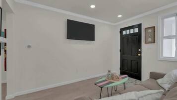 Un salon avec un canapé beige, une petite table ornée de quelques objets décoratifs, une porte noire et un téléviseur à écran plat fixé au mur.