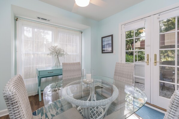 Una habitación con una mesa redonda de vidrio, sillas de mimbre, una mesita auxiliar azul y una puerta blanca con paneles de vidrio.