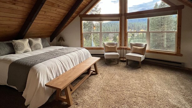 Un dormitorio con una cama grande, un banco, dos sillones y ventanas amplias que ofrecen vistas a un paisaje boscoso.