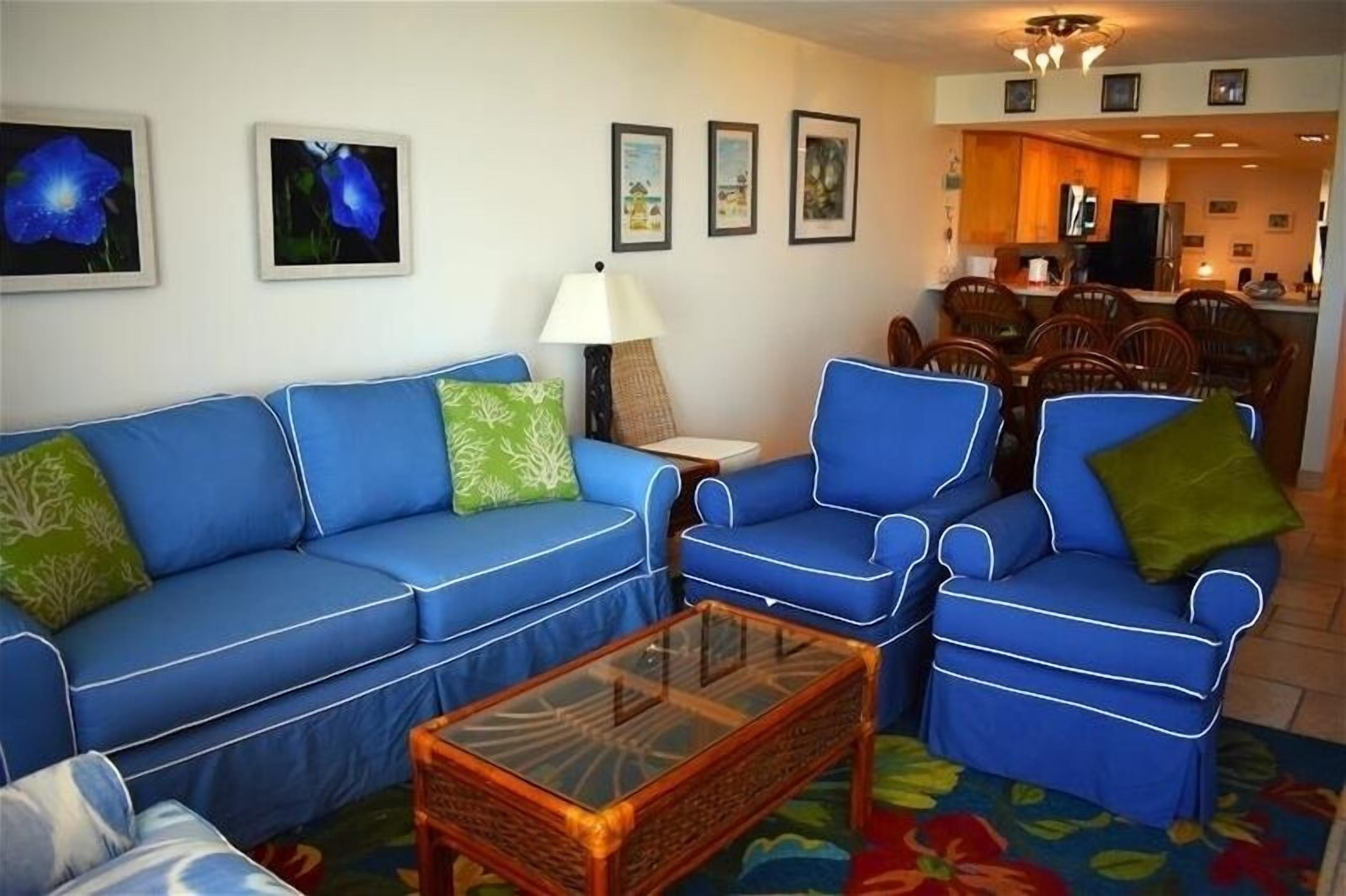 Un salon avec un canapé bleu, des fauteuils assortis, une table basse en verre et un coin repas comprenant des chaises et une table en bois.