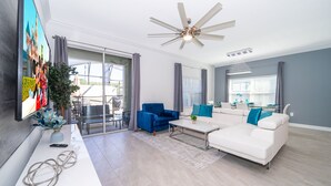 A modern living room with a white sofa, blue armchair, and a coffee table. There is a ceiling fan, a TV mounted on the wall, and a large window with curtains. - RH4415 Townhouse 5/4 Private Pool & Free Waterpark (Kissimmee)