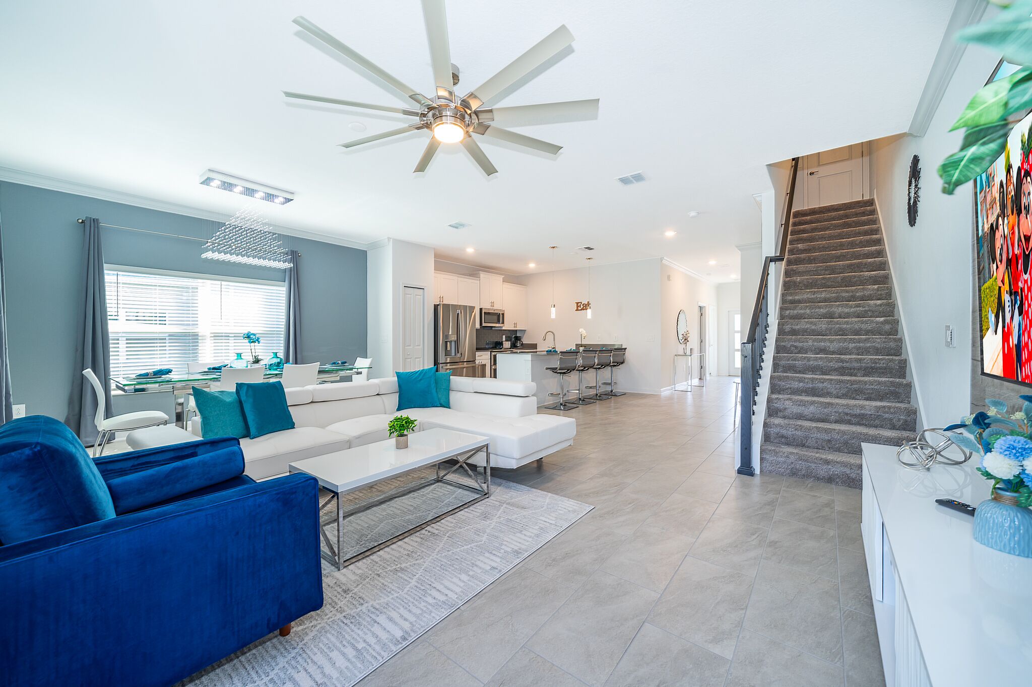 Ein modernes Wohnzimmer mit einem weißen Sofa, einem blauen Sessel und einem Couchtisch. Im Hintergrund ist eine Küche erkennbar, und eine Treppe führt zu einer weiteren Ebene.