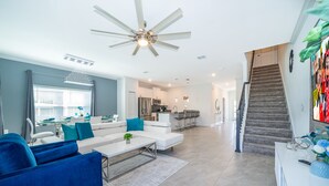 A modern living room with a white sofa, blue armchair, and a coffee table. A kitchen is visible in the background, and a staircase leads to another level. - RH4415 Townhouse 5/4 Private Pool & Free Waterpark (Kissimmee)