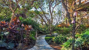Un sentier de pierres serpente à travers un jardin luxuriant, où poussent différentes plantes et arbres, et mène à un bâtiment surmonté d’un toit en tuiles.