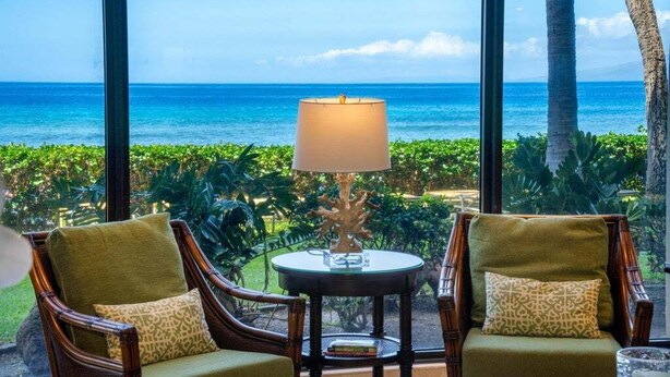 Une pièce avec vue sur l’océan, deux fauteuils avec des coussins, une table avec une lampe et un décor de plantes tropicales.