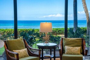Une pièce avec vue sur l’océan, deux fauteuils avec des coussins, une table avec une lampe et un décor de plantes tropicales.