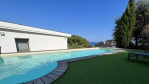 A swimming pool with a wooden deck and artificial grass, surrounded by a white building and trees. - Ground floor villa with private pool (Cavalaire-sur-Mer)