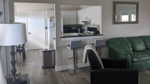 Eine moderne Küche mit weißen Schränken, einer Mikrowelle und einer Kaffeemaschine. Im Wohnbereich befindet sich eine grüne Ledersofa und ein kleiner Tisch mit einer Lampe.
