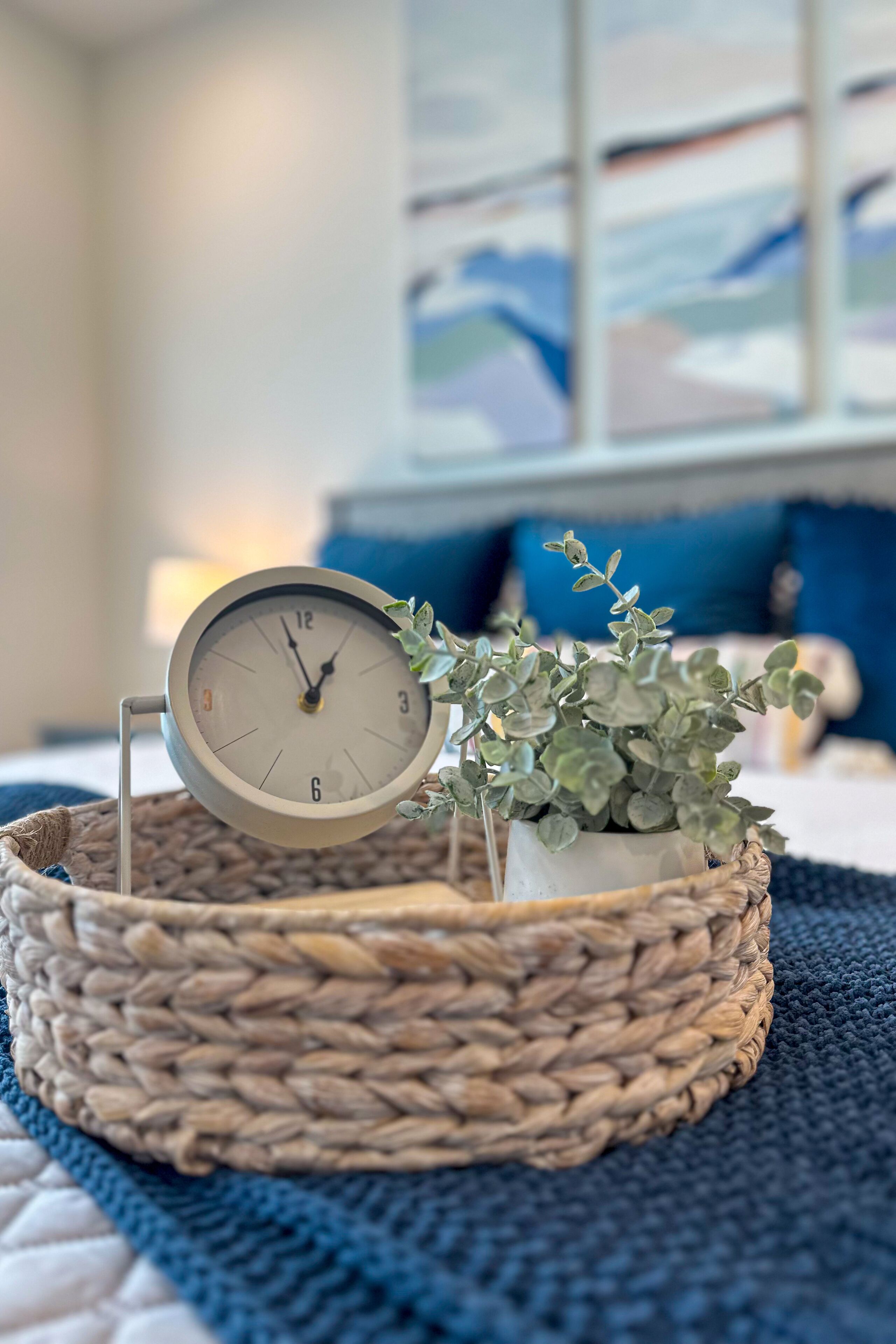Un panier tressé contenant une horloge et une petite plante en pot, posé sur un lit recouvert d’une couverture bleue.