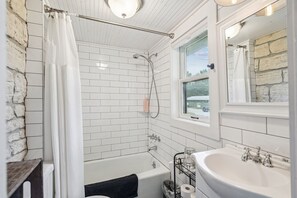 Une salle de bain aux murs recouverts de carreaux blancs, avec une baignoire, une douche, un lavabo et une fenêtre donnant sur une maison et des arbres.