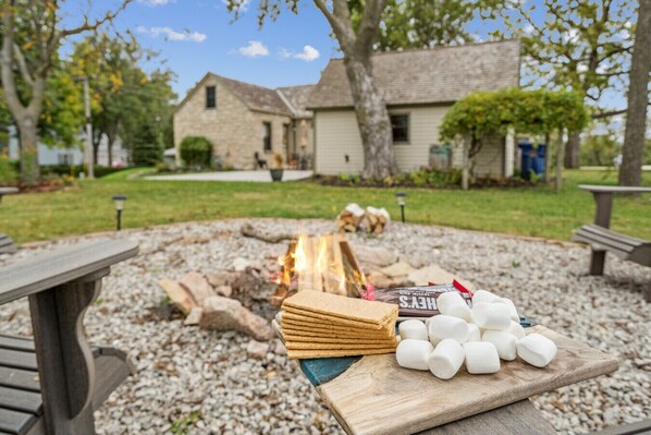 A picnic setup with a fire, marshmallows, and chocolate bars on a wooden table in a park area with stone buildings in the background. - The Rock House • Historic Stone Cottage • Walk to Central (Pella)
