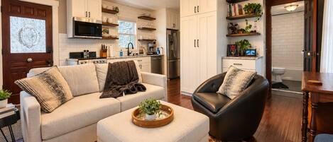 A modern living room with a sofa, ottoman, and leather chair, a kitchen in the background, and a dining area with a wooden table.