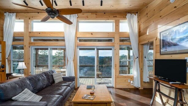 Interior acogedor de una cabaña con sofá de cuero, mesa de centro de madera y ventilador de techo. Amplias ventanas ofrecen vistas del exterior.