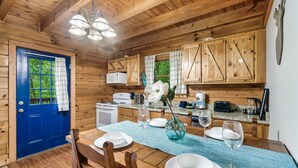 Interior de una cabaña de madera con una mesa de comedor preparada para cuatro, armarios de madera, una puerta azul y una zona de cocina con microondas y cafetera.