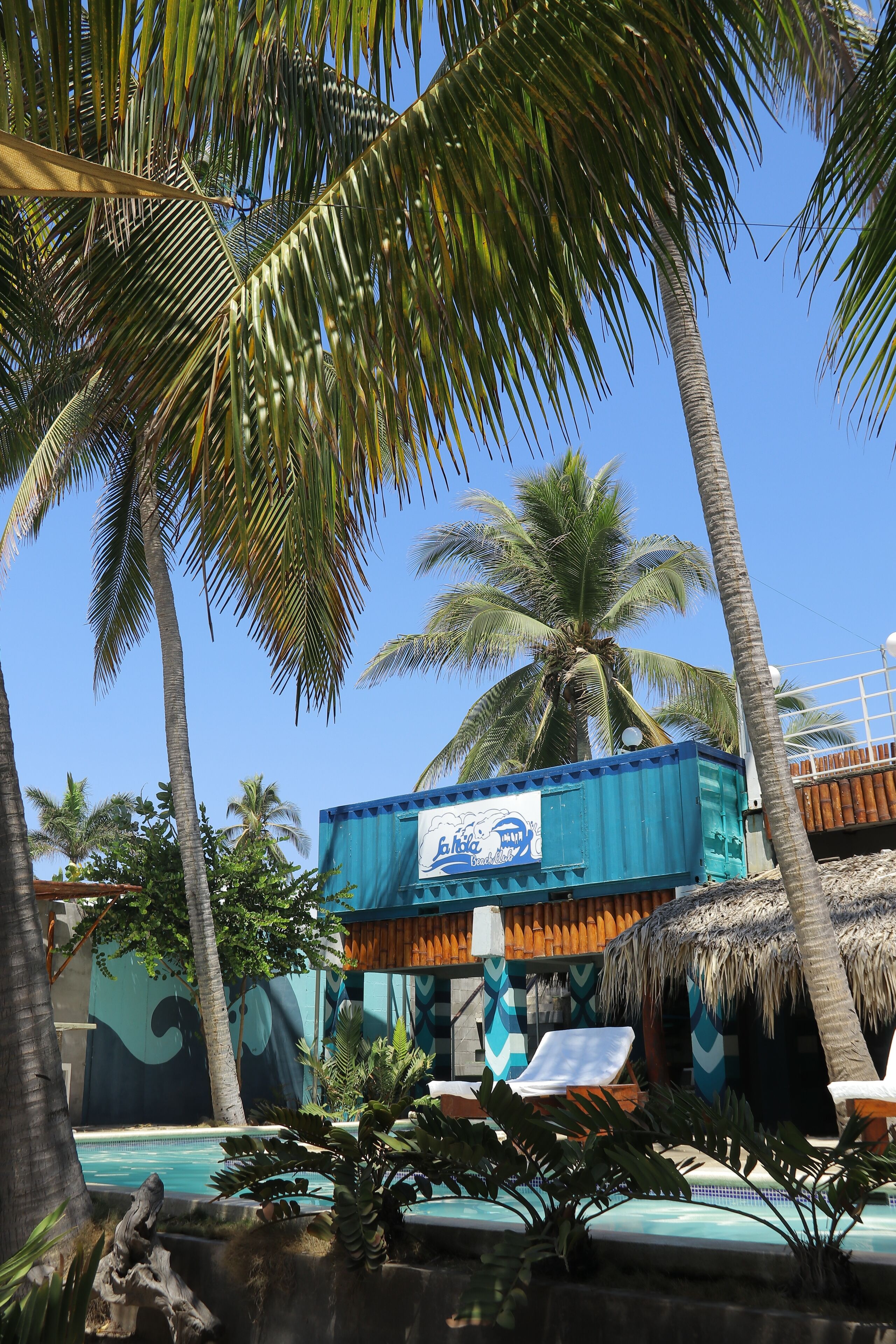 Zona de piscina con sillones de descanso, palmeras y un edificio con un letrero azul.
