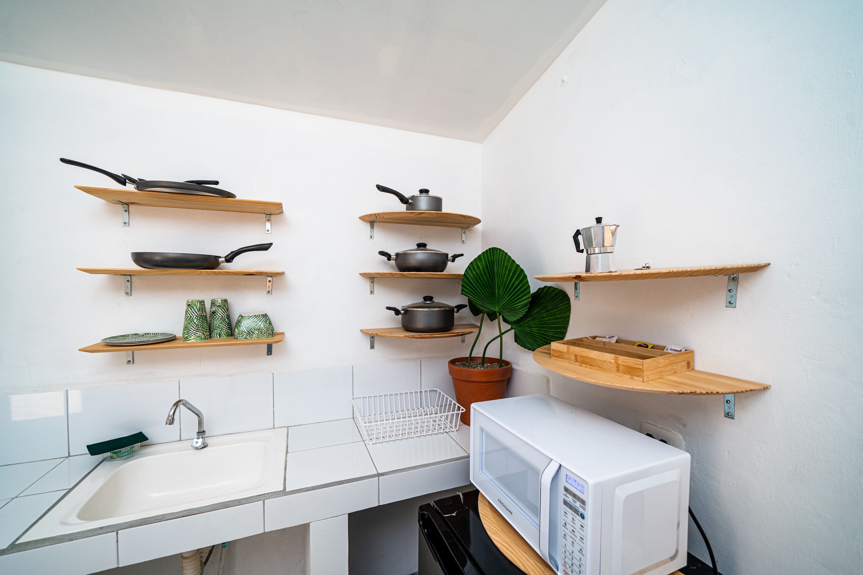 Une cuisine avec des carreaux blancs, un évier, un four à micro-ondes et des étagères où sont rangés les ustensiles de cuisine et une plante en pot.
