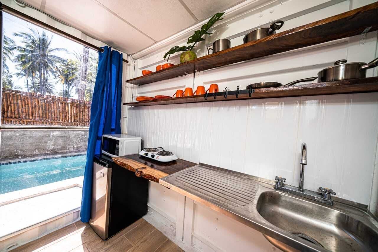 Een compacte keuken met een inbouwfornuis, een spoelbak en boven de aanrechtplaten staan keukengerei op planken.