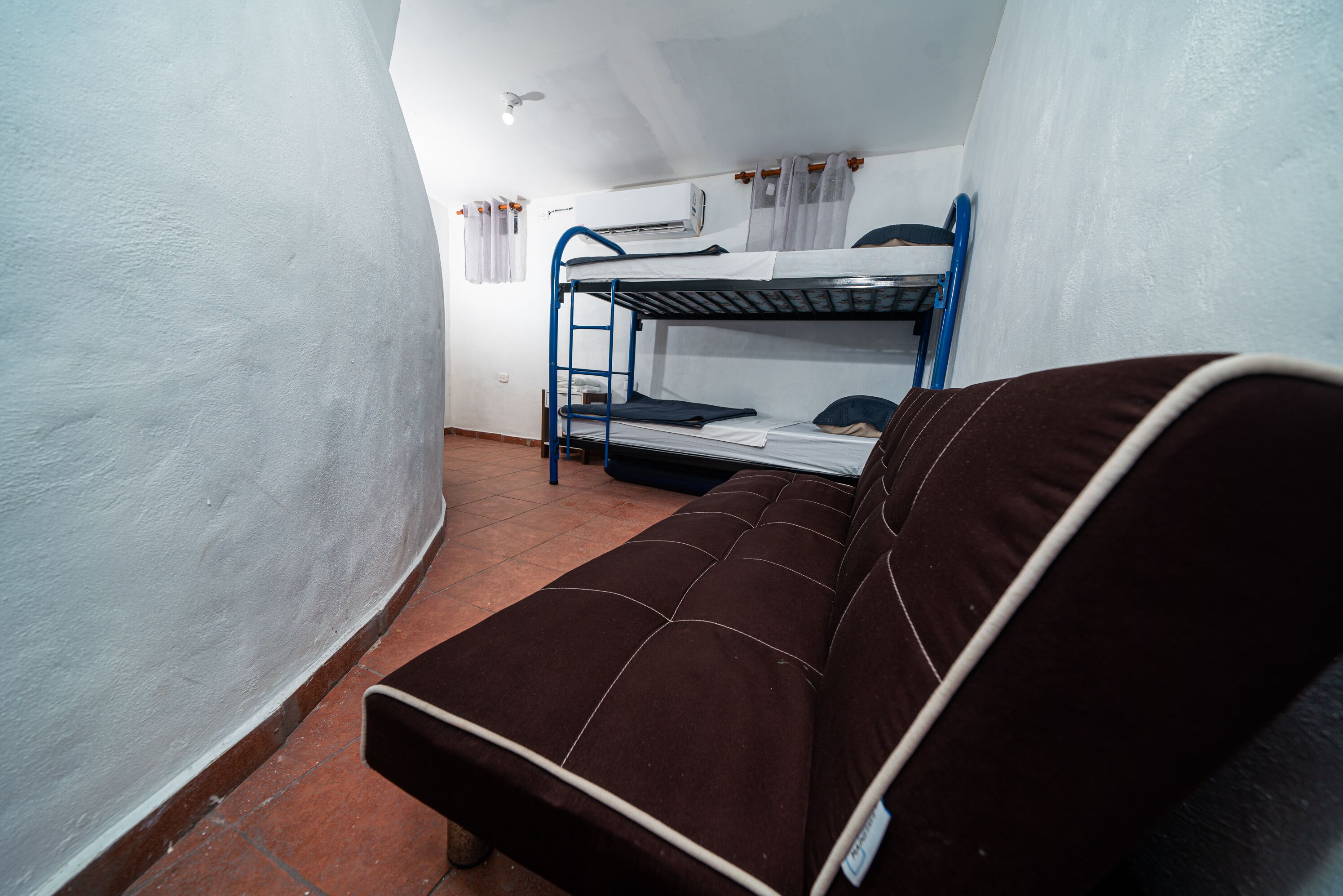 A dormitory room with a bunk bed, a single bed, a small table, and a ceiling fan.