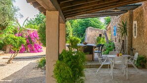 A covered outdoor patio with a table and chairs, surrounded by greenery and a stone wall.
