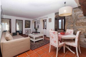 Un soggiorno con divano, tavolino e poltrone floreali. Si nota un tavolo da pranzo con sedie e un lampadario. La stanza presenta una parete in pietra e pavimento piastrellato.