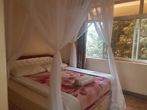A bed with a canopy, a window with a view of trees, and a bedside table. - Kilimanjaro Star Hotel (Moshi)