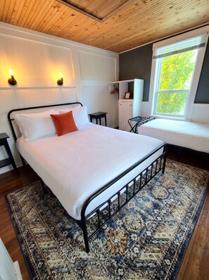 Un dormitorio con cama, mesita de noche, lámpara colgante, ventana con vistas a la vegetación y alfombra estampada.