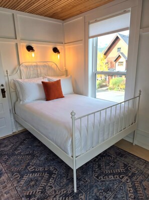 Een wit metalen bed met hoofdeinde, twee wandlampen, een raam met uitzicht op huizen en een kamerbreed tapijt met een patroon.