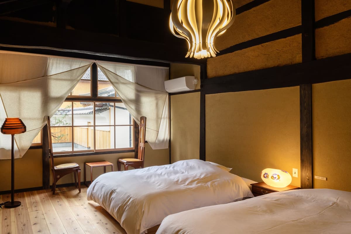 Een traditionele Japanse kamer met tatami vloerbedekking, een laag houten bed, een hanglamp en uitzicht op een tuin door een schuifdeur.