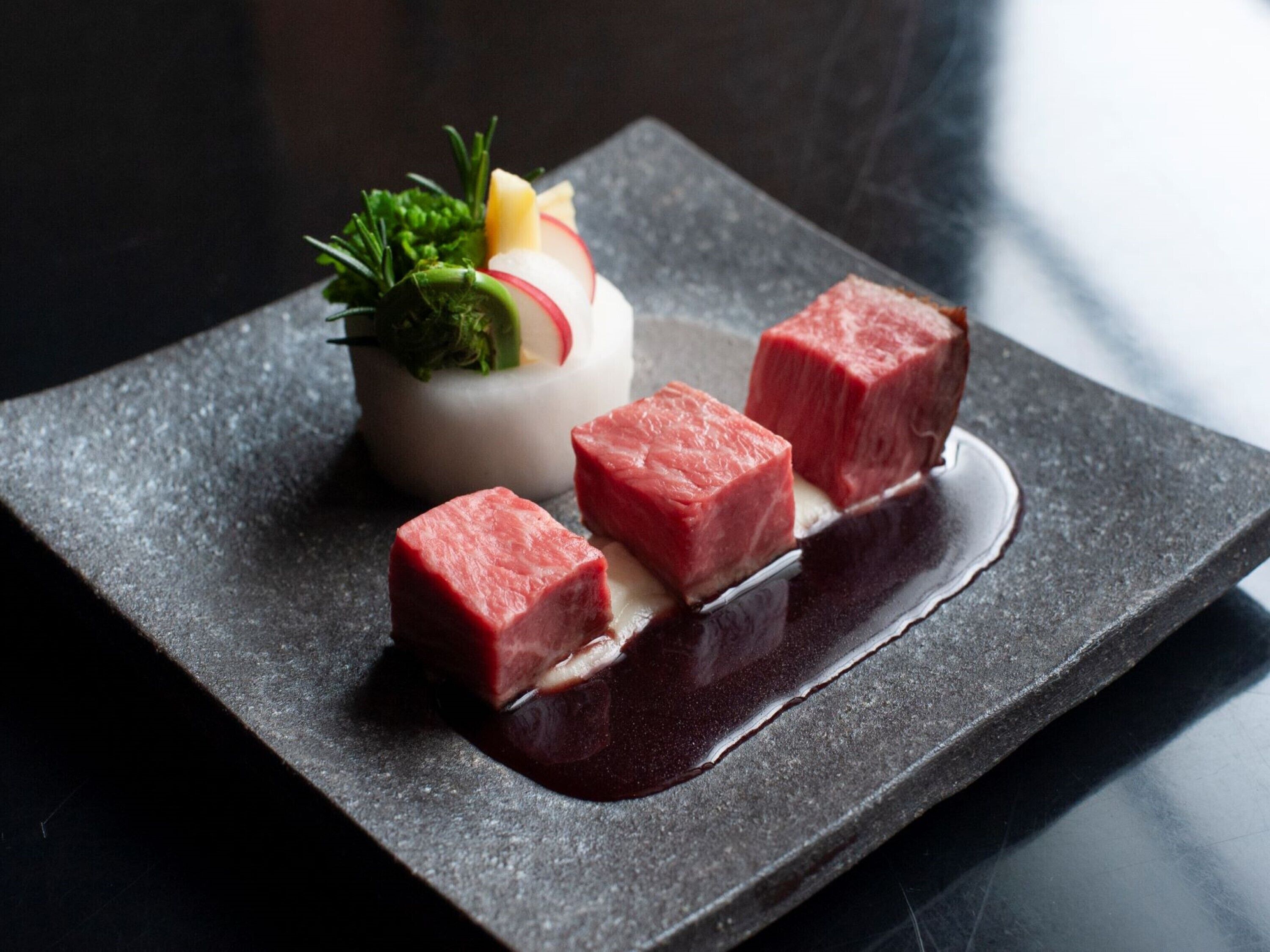 A square plate with three cubes of raw meat and a small bowl of vegetables on a dark surface.