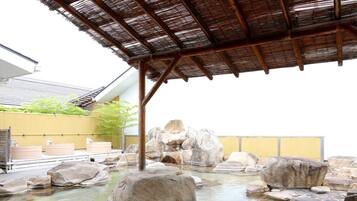 A traditional Japanese onsen with a wooden structure overhead, large rocks in the water, and a yellow fence in the background.