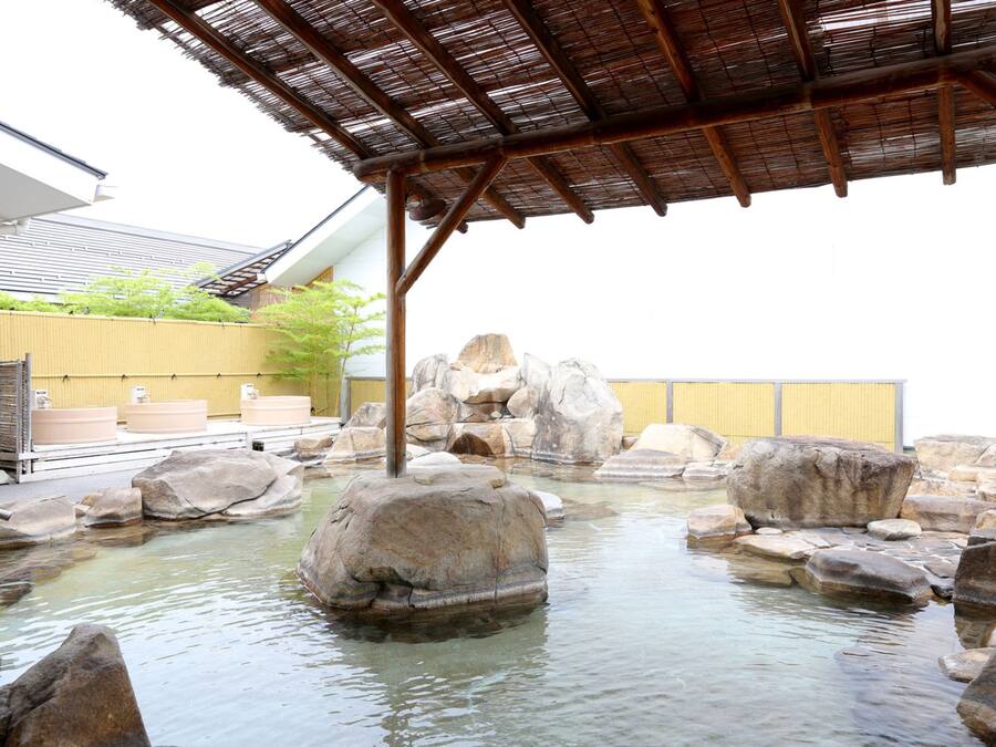 A traditional Japanese onsen with a wooden structure overhead, large rocks in the water, and a yellow fence in the background.