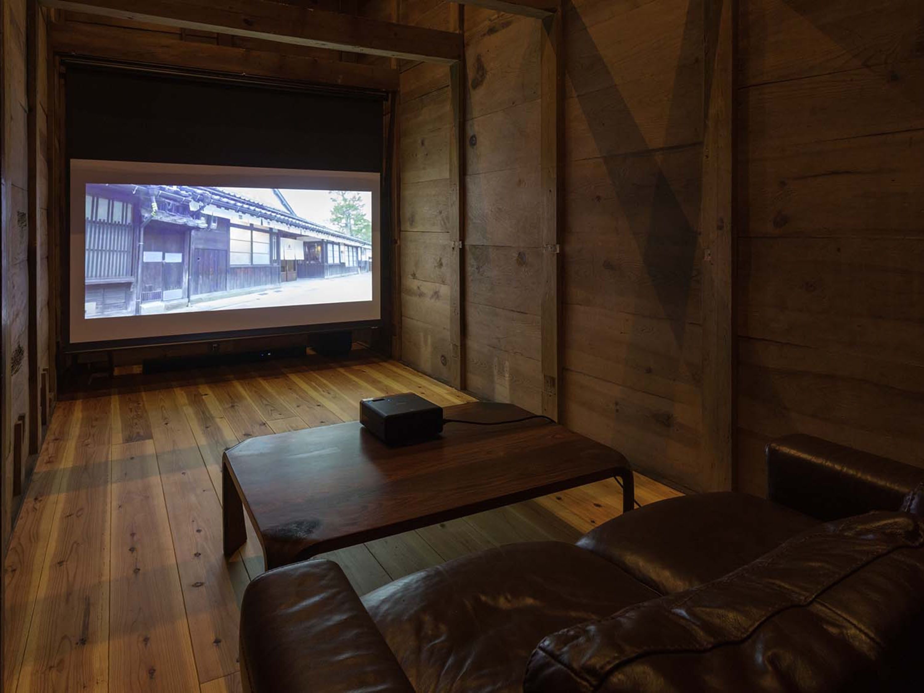 A cozy room with a wooden floor, a leather couch, a small table, and a large screen displaying an image of a building.