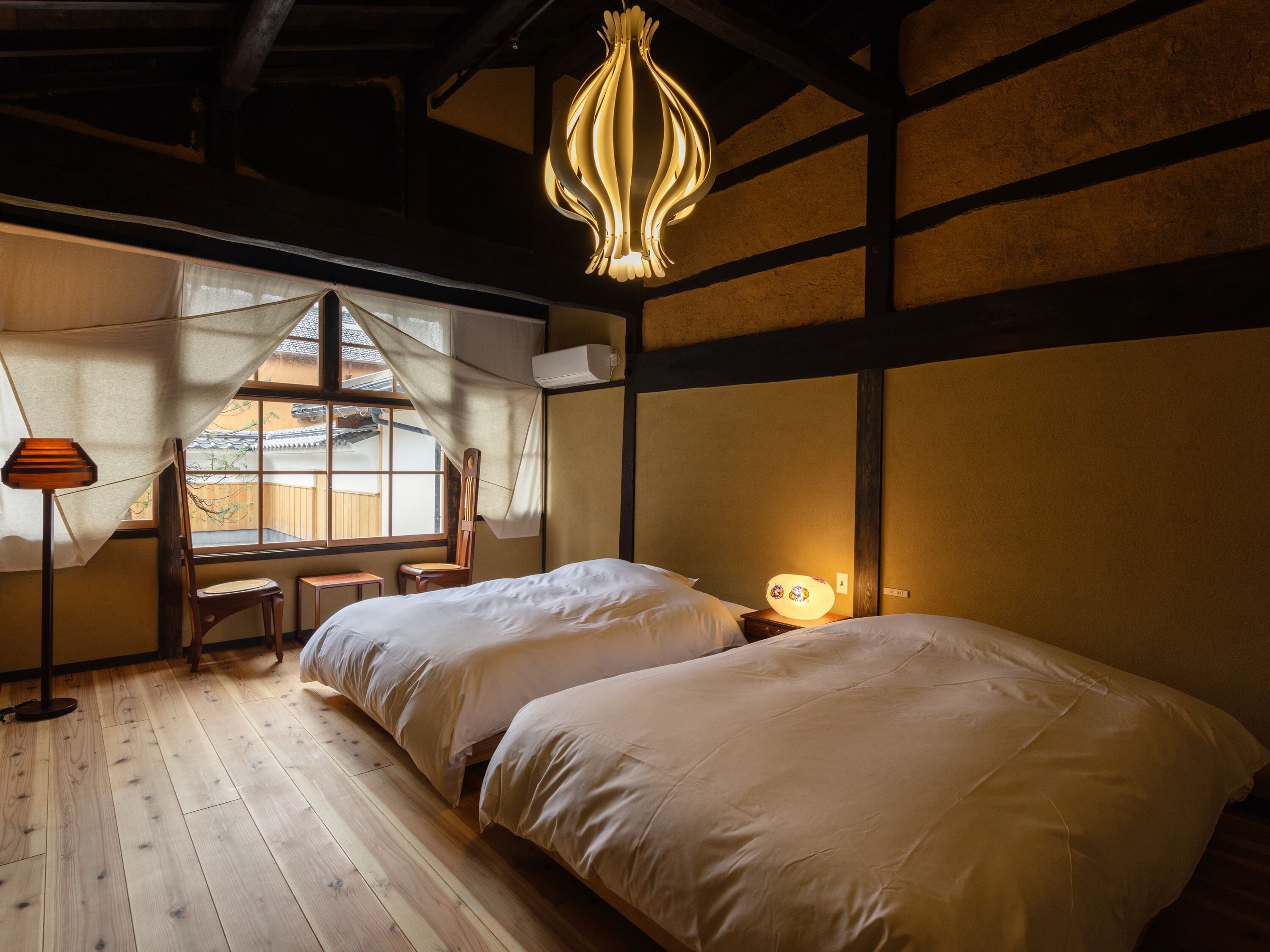 A traditional Japanese-style room with two beds, a wooden floor, a hanging light fixture, and a view of a traditional building outside.