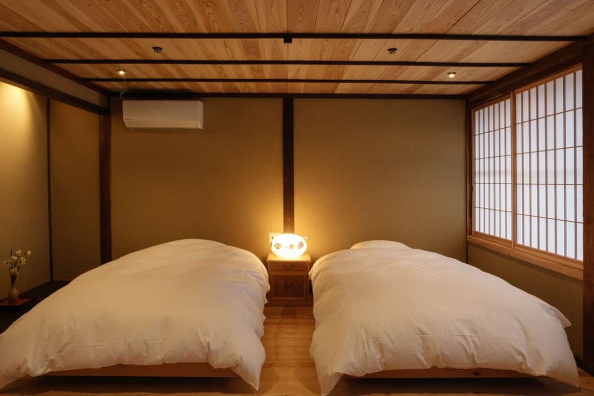 Een kamer met twee eenpersoonsbedden, houten vloer, een houten plafond met zichtbare balken, een wandairconditioner en een raam met verticale jaloezieën.
