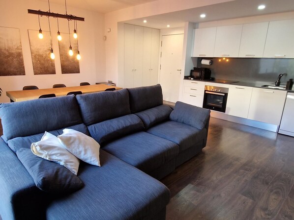 A modern living room with a grey sofa, a dining table with chairs, and a kitchen area with white cabinets and appliances.