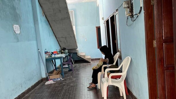 Un pasillo con suelo de baldosas, una persona sentada en una silla de plástico y una puerta a la derecha.