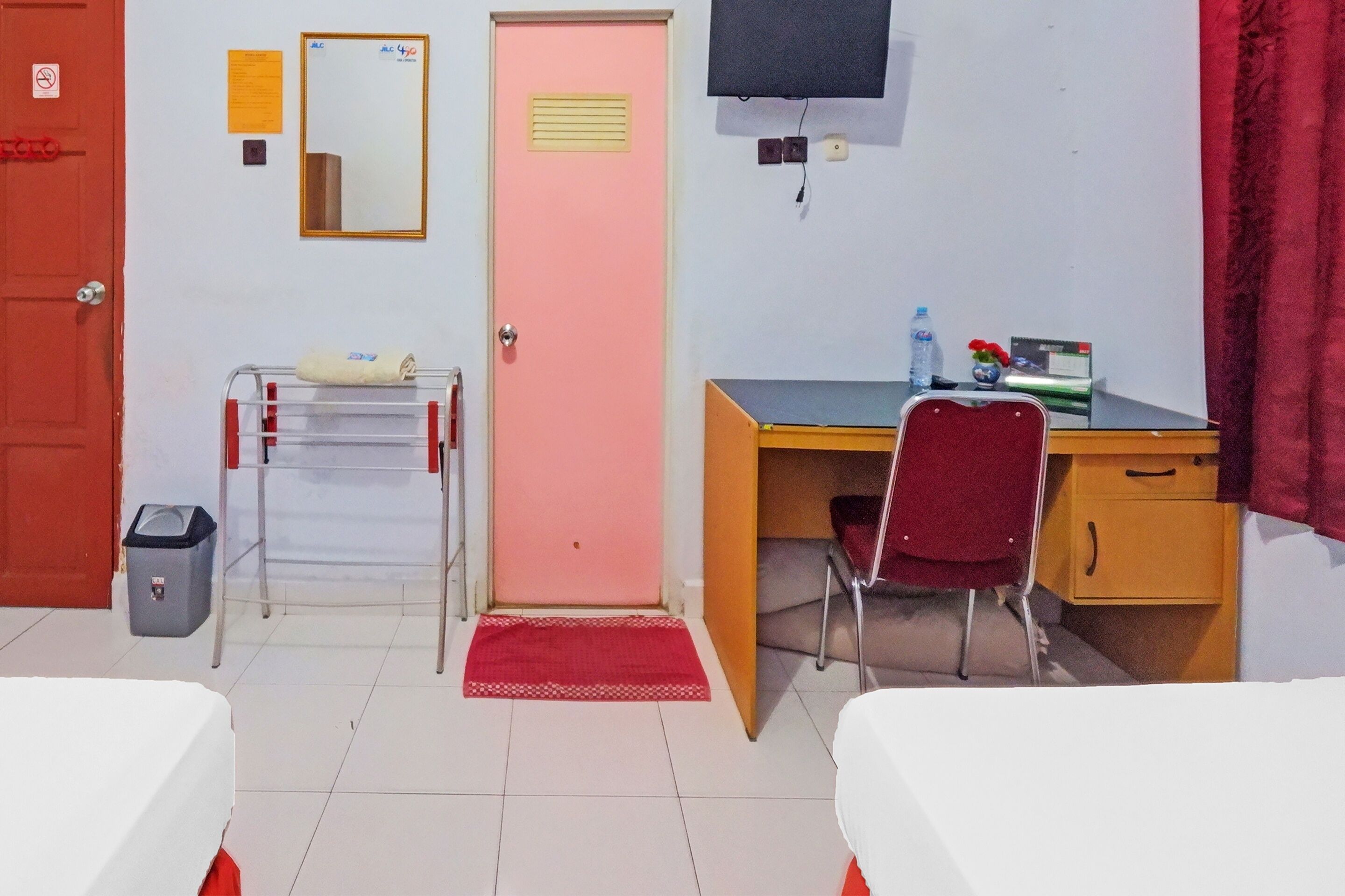 Une chambre d’hôtel avec une porte rose, un bureau avec une chaise, un lit, une poubelle et un petit tapis.