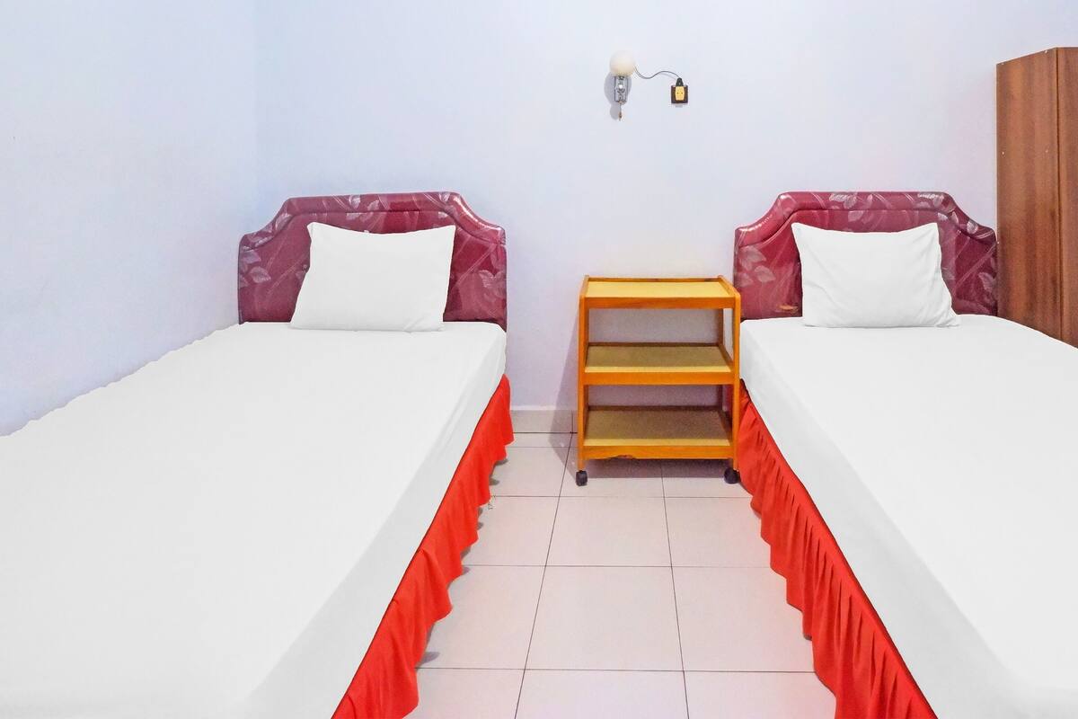 A room with two beds, each with a red skirted base, a wooden nightstand between them, and a wall-mounted light fixture.
