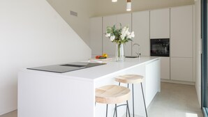 Uma cozinha moderna com armários brancos, uma ilha central com banquetas, iluminação pendente e um vaso com flores.