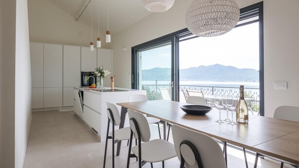 Uma cozinha moderna com armários brancos, uma área de jantar com mesa e cadeiras de madeira, e uma grande janela que oferece vista para montanhas e um lago.