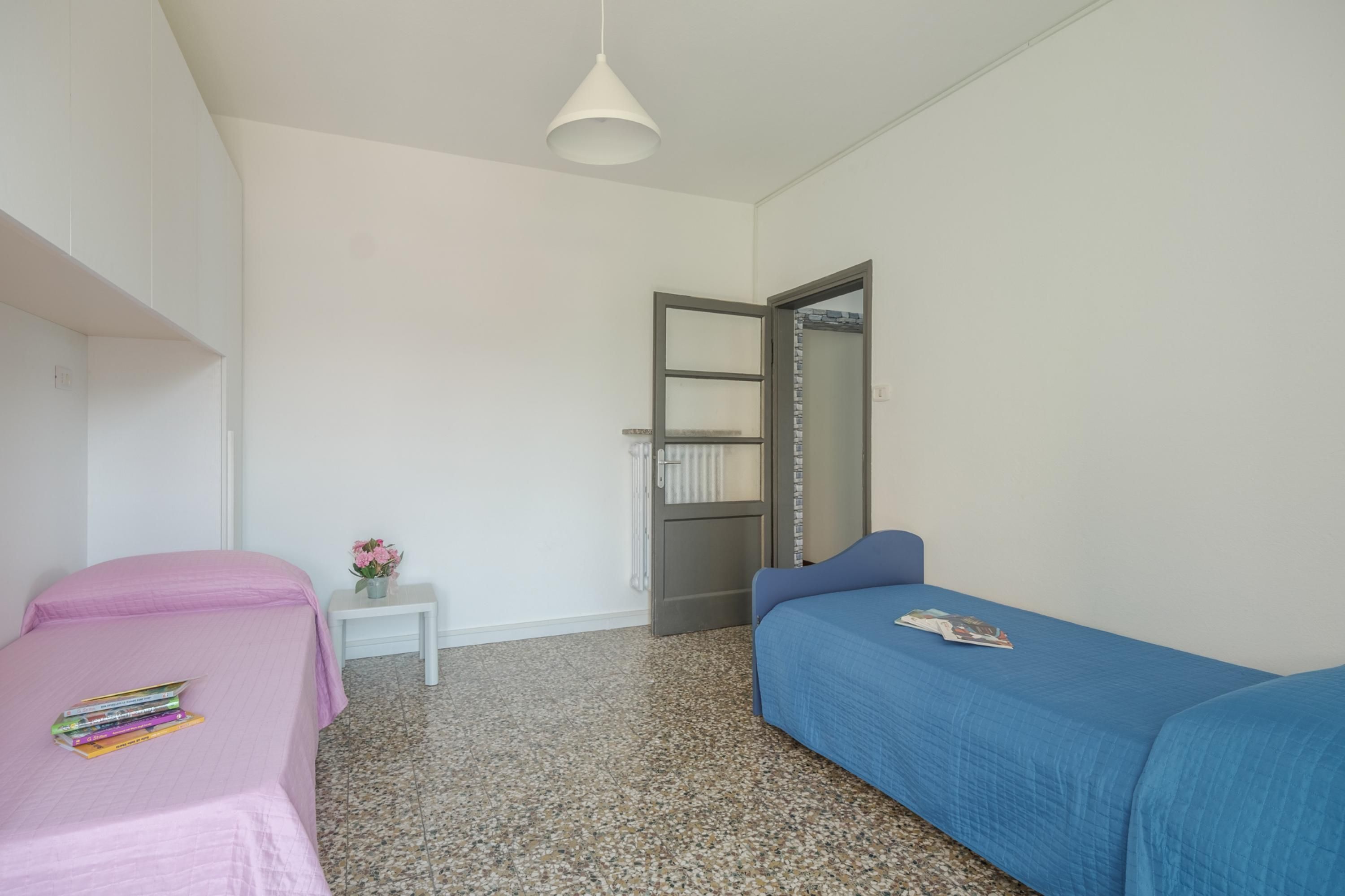 A bedroom with two beds, a small bedside table with a vase, and a door leading to a bathroom.