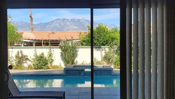 Vista desde una habitación con puerta corrediza de vidrio hacia una piscina, un área de patio y montañas al fondo.