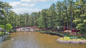 A serene lakeside setting with a wooden dock, surrounded by tall pine trees and a small building with a red roof. - Lake view 2 bedroom House on Lake Delton Resort. Beaches. Pool. Playground! (Lake Delton)
