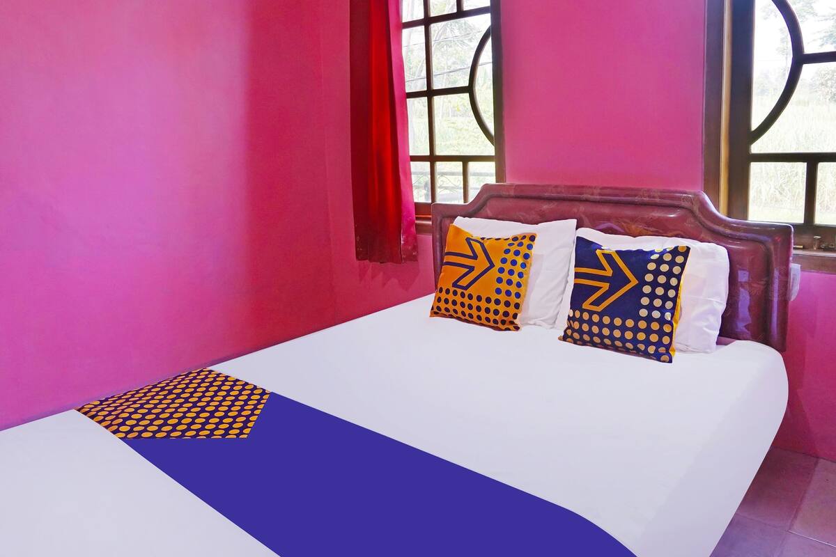 Un lit recouvert d’une couvre-lit blanche et bleue, deux oreillers à motifs géométriques et une fenêtre avec un rideau rouge.