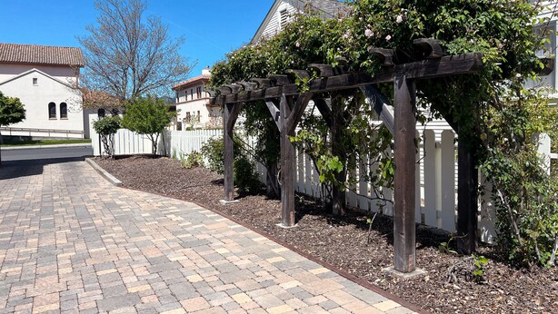 Un camino pavimentado que conduce a una pérgola de madera con plantas trepadoras, rodeada por una cerca blanca y casas con techos de tejas rojas.