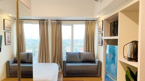 Ein modernes Hotelzimmer mit einem Bett, zwei Sofas, einem großen Fenster mit Vorhängen, einer Küchenzeile und Blick auf die Stadt.