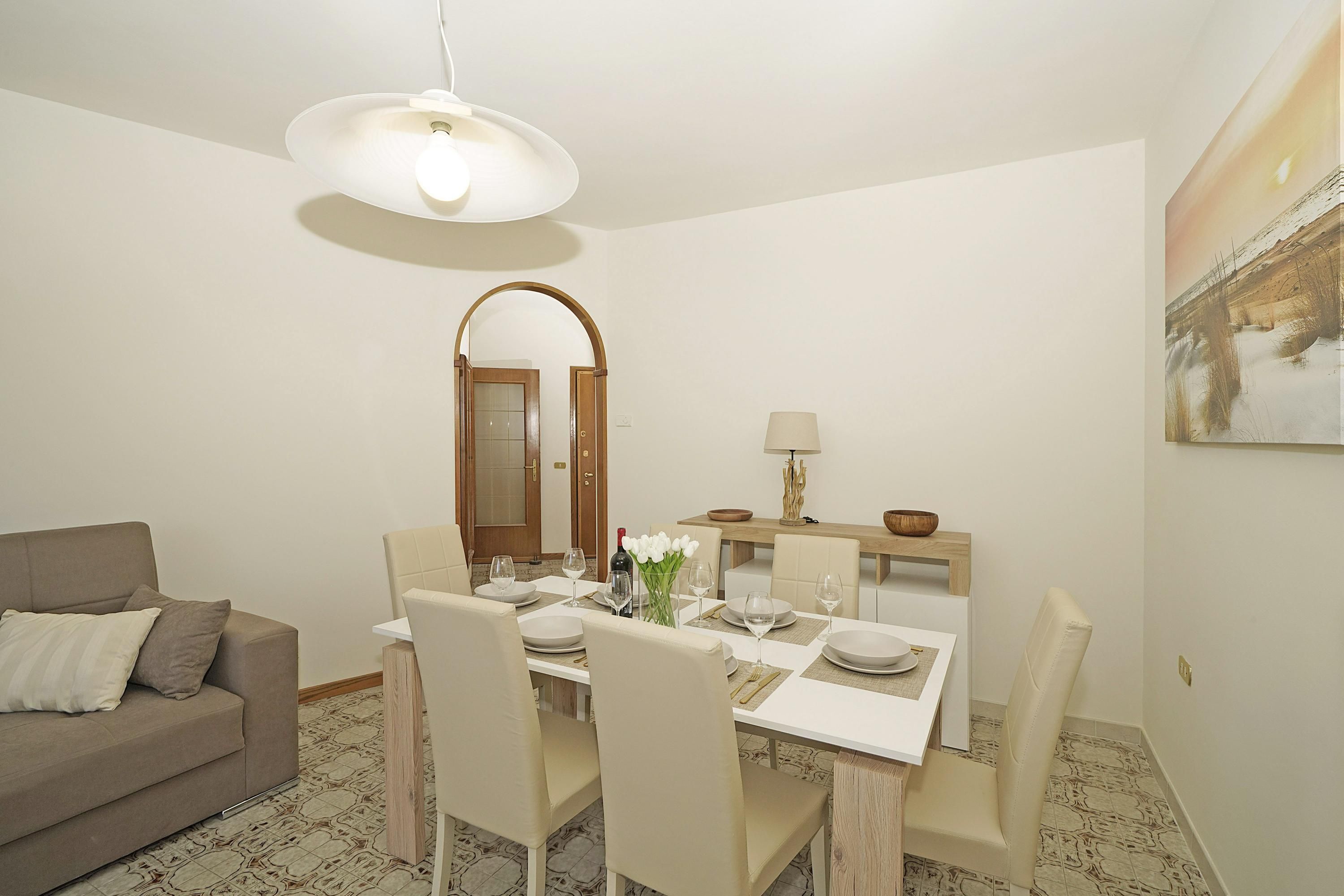 Uma sala de jantar com uma mesa posta para quatro pessoas, um sofá, um espelho e um quadro na parede.