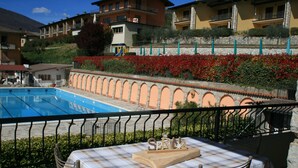 A poolside area with a checkered table and chairs, a sign reading "SPRING," and a view of residential buildings and a playground in the background. - Appartamento Anna 701 (Tignale)